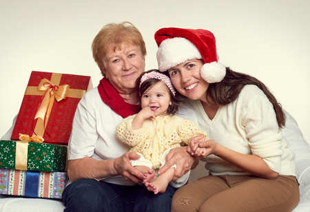 Grandma, daughter and granddaughter dressed in santa hat with gift boxes sit on sofa, white background. New year eve and christmas holiday concept.の写真素材