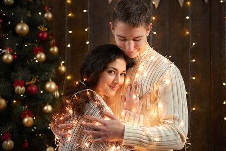 young couple together in christmas lights and decoration, dressed in white, fir tree on dark wooden background, romantic evening, winter holiday conceptの写真素材