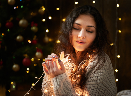 dreaming girl portrait with handful of christmas lights, closed eyes, dressed in white sweater, dark wooden background, winter holiday conceptの写真素材