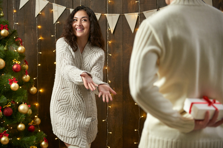 Happy couple give gifts in christmas decoration, sitting on floor in dark wooden interior with lights. Romantic evening and love concept. New year holiday.の写真素材