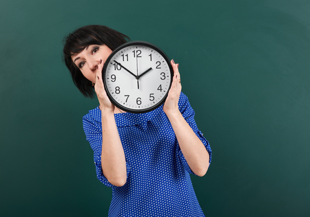 woman with big clock posing by chalk board, time and education concept, green background, studio...