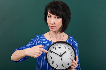 woman pointing finger at the watch, posing by chalk board, time and education concept, green background, studio shotの写真素材