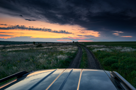 the car rides on a dirt road in the field, beautiful sunset with wild grass, sunlight and dark cloudsの写真素材