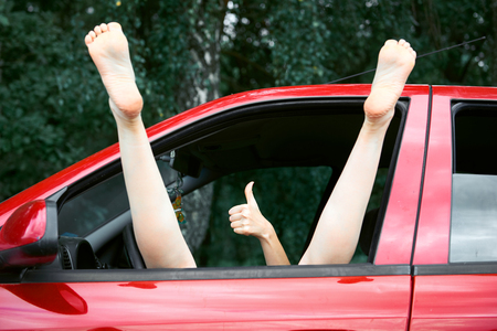 young woman driver resting in a red car, put her feet on the car window and gesturing, happy travel conceptの写真素材