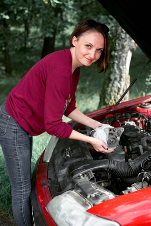 a young girl stands at a broken car and holds a bad spare part, an electric generator fixed with bandage, happy emotion about the end of the repairの写真素材