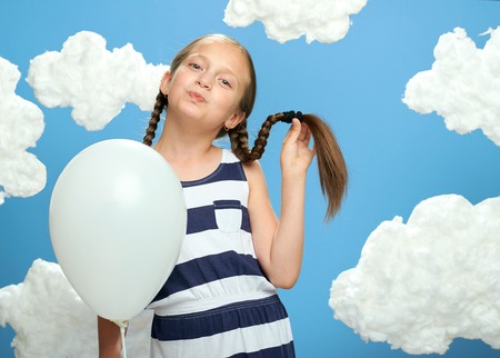 girl dressed in striped dress posing on a blue background with cotton clouds, white air balloon, the concept of summer and happinessの写真素材