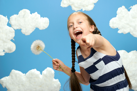 girl has a big dandelion in her hands, dressed in striped dress, posing on a blue background with cotton clouds, the concept of summer, holiday and happinessの写真素材