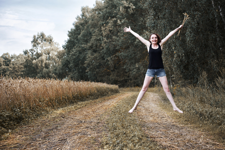 young girl jumping barefoot on the ground road through field and forest, the concept of summer and travelの写真素材