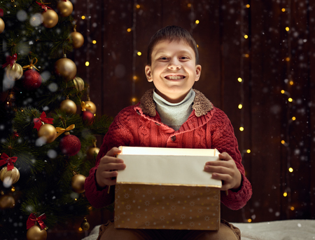 child boy sitting with gift box near christmas decorated fir tree, dark wooden backgroundの写真素材
