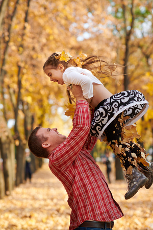 Father throws the daughter high up in autumn city park. They posing, smiling, playing. Bright yellow trees.の写真素材