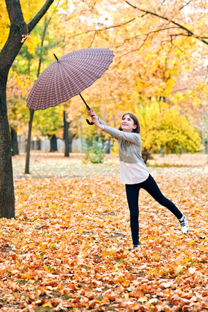 Teen girl with umbrella posing in autumn park. Bright yellow leaves and trees. She imitates the wind.の写真素材