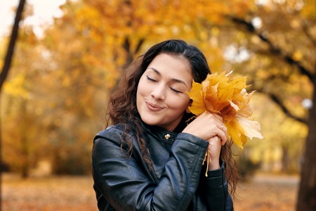 Pretty woman is posing with bunch of maple's leaves in autumn park. She's closing eyes and dreaming....