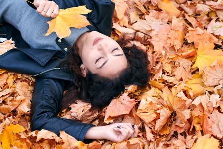 Pretty woman is lying in autumn park on fallen leaves. Beautiful landscape at fall season.の写真素材