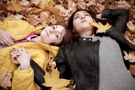 Pretty woman and teen girl are posing in autumn park. They are lying on fallen leaves. Beautiful landscape at fall season.の写真素材