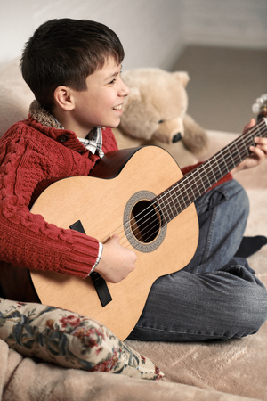 boy plays with an acoustic guitar, sits on the sofa with a bear toyの写真素材
