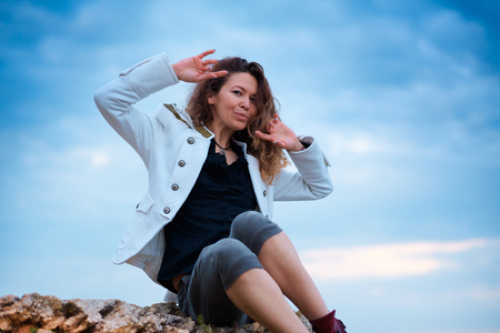 fashionable girl dressed in white jacket and wide trousers posing at sunset sky background, sits on the stoneの写真素材