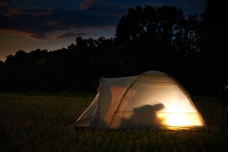 Traveling and camping concept - camp tent at night under a sky full of stars. Orange illuminated tent with a person inside. Beautiful nature - field, forest, plain. Moon and moonlightの写真素材