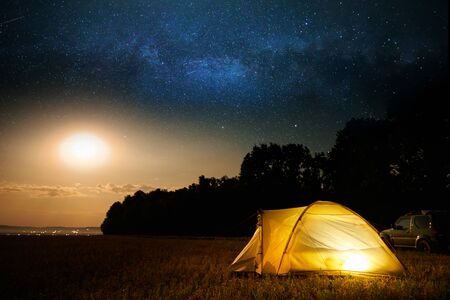 Traveling and camping concept - camp tent at night under a sky full of stars. Orange illuminated tent and car. Beautiful nature - field, forest, plain. Moon and moonlightの写真素材