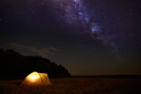 Traveling and camping concept - camp tent at night under a sky full of stars. Orange illuminated...