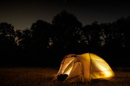 Traveling and camping concept - camp tent at night under a sky full of stars. Orange illuminated tent. Beautiful nature - field, forest, plain. Moon and moonlightの写真素材