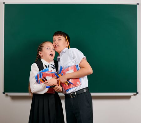 Emotional portrait of a schoolboy and schoolgirl posing with exercise books, pens, pencils and other school supplies on blackboard background - back to school and education conceptの写真素材