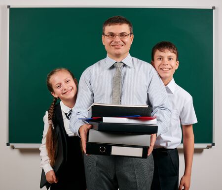 Portrait of a teacher dressed in business suit with folders, documents and briefcase, posing with schoolboy and schoolgirl at blackboard background - learning and education conceptの写真素材