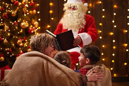 Santa Claus reading book for family. Mother and children sitting indoor near decorated xmas tree with lights - Merry Christmas and Happy Holidays!の写真素材