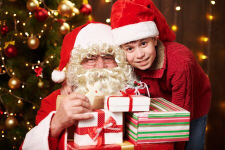 Santa Claus and santa helper boy posing with many gifts, sitting indoor near decorated xmas tree with lights - Merry Christmas and Happy Holidays!の写真素材