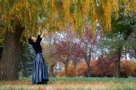 Beautiful elegant woman standing and posing near big yellow tree in a city park in autumnの写真素材