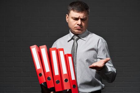 Very busy businessman closeup portrait, posing with red folders, overworking concept, dark wall backgroundの写真素材
