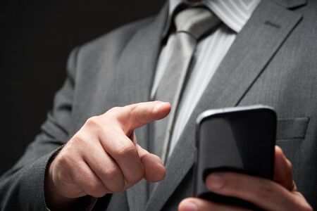 businessman portrait dressed in gray suit, dialing by smartphone, dark wall background