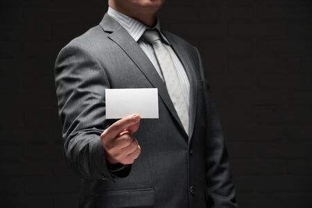 white blank business card closeup in businessman hand, gray suit, dark wall backgroundの写真素材