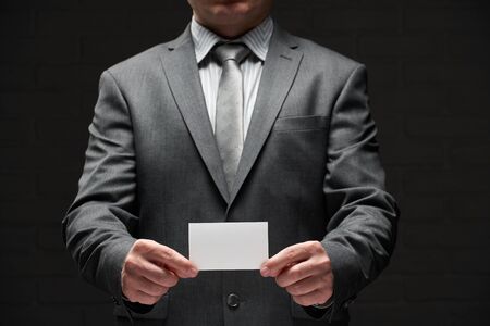 white blank business card closeup in businessman hand, gray suit, dark wall backgroundの写真素材