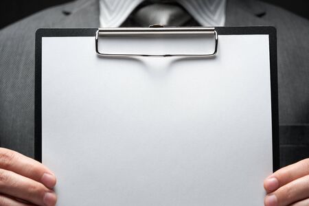 white blank paper sheet closeup in businessman hand, gray suit, dark wall background, blank space...