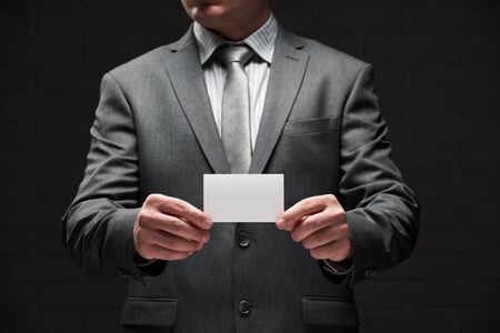 white blank business card closeup in businessman hand, gray suit, dark wall backgroundの写真素材