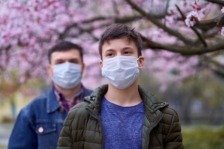 father and son with a face mask are in the city outdoor, blooming trees, spring season, flowering time - concept of allergies and health protection from dusty airの写真素材