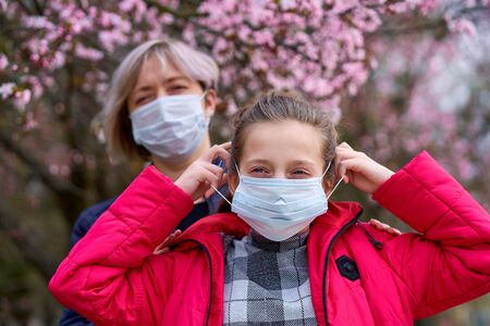 mother and daughter with a face mask are in the city outdoor, blooming trees, spring season, flowering time - concept of allergies and health protection from dusty airの写真素材