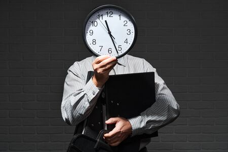Businessman closeup portrait, he stands and shows the clock in front of his head, time and schedule concept, dark wall backgroundの写真素材