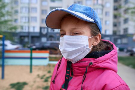 a girl sitting on a seesaw on playground near high-rise buildings with apartments, a medical mask on her face protects against viruses and dustの写真素材