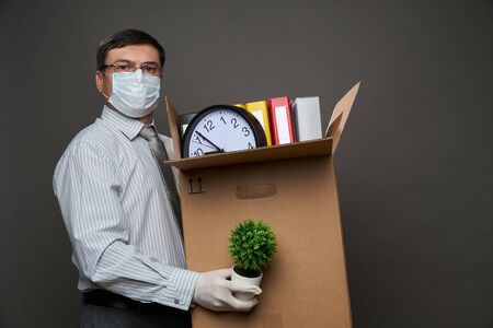 a man dressed as a businessman holds a box with office stuff, documents, posing in studio on gray...