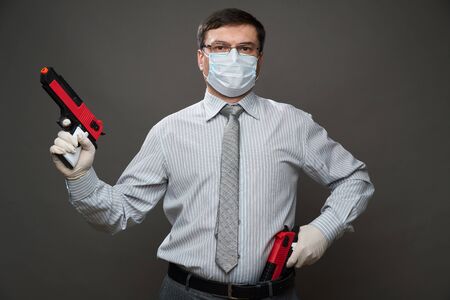 a man dressed as a businessman, posing in studio on gray background, medical face mask and protective gloves, gun, glasses, shirt and tie - concept of quarantine and antivirus protectionの写真素材