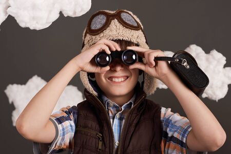 a boy plays with binoculars and dreams of becoming a pilot, dressed in a retro style jacket and helmet with glasses, clouds of cotton wool, gray background, tinted in brownの写真素材