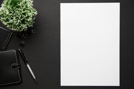 Top view of modern black office desk with blank paper sheet, notebook, pen and a lot of things. Flat...
