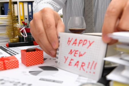 Business and holiday concept - businessman celebrating in office decorated with Christmas tree, candies and other new year decoration. Shows a notepad with happy new year textの写真素材