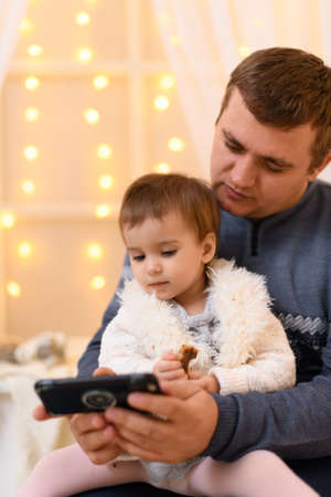 father and baby use smartphone, they play and watch device screen, home interior decorated with...