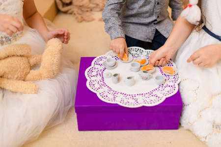 children drink tea, eat cookies and play with the toy dishes in home interior decorated with holiday lights and giftsの写真素材