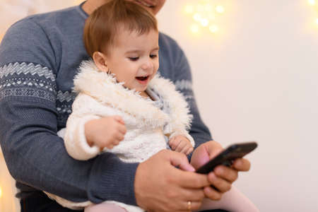 father and baby use smartphone, they play and watch device screen, home interior decorated with holiday lights and giftsの写真素材