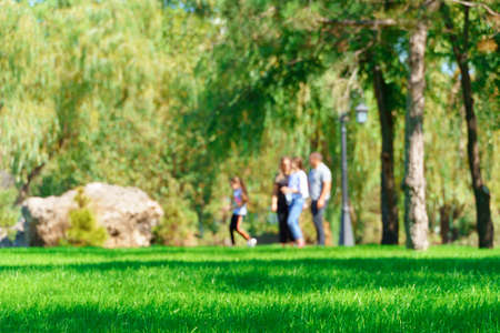 people walking and children playing in a city park on a summer day, green lawns with grass and trees, paths and benches, bright sunlight and shadowsの写真素材
