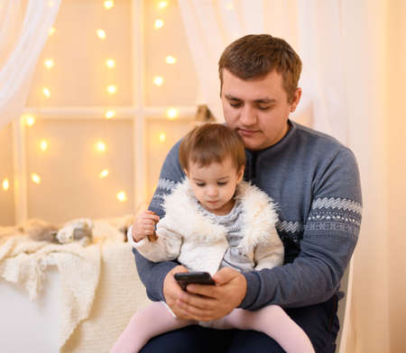 father and baby use smartphone, they play and watch device screen, home interior decorated with holiday lights and giftsの写真素材