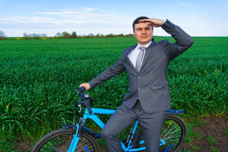 businessman with a bicycle poses in a wheaten field, green grass and blue sky as backgroundの写真素材